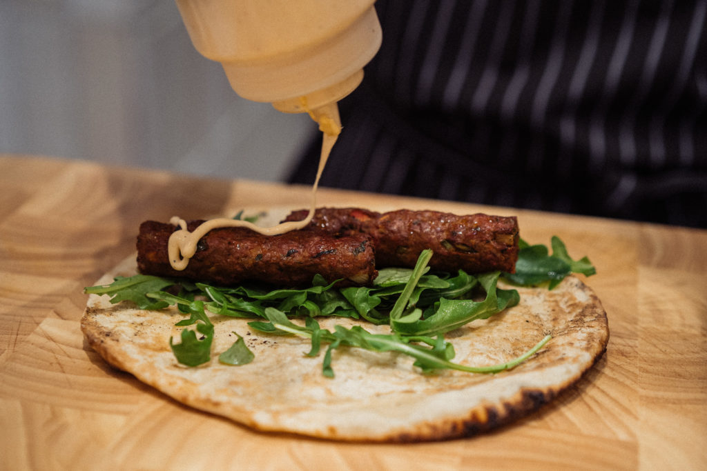 Naan bread with leafy salad on top, with two spicy lamb kebabs and sauce being drizzled on top. 