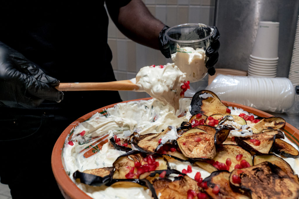 Man transferring yoghurt sauce to clear plastic container from large dish of prepared yoghurt. Yoghurt decorated with sliced roasted aubergine and sprinkled with pomegranate seeds. 