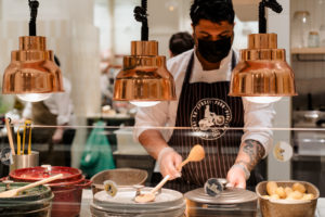 Chef wearing Dabbawal apron working behind service counter with utensil in his hand