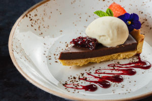 Slice of home-made chocolate tart served on a plate, with a scoop of ice-cream and flower and fruit decoration 