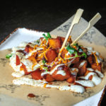 Cubes of sweet potato piled on plate, covered in sauces and garnishes, with two forks to eat the dish with