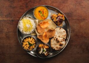Bird’s eye view of round silver platter, with 7 dishes arranged in a circle, items include small papads, yellow lentils, curry, rice and bread