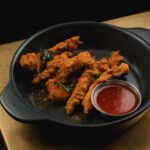 Chicken battered fritters arranged in black dish, accompanied by a small dish of sweet chilli sauce