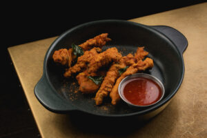 Chicken battered fritters arranged in black dish, accompanied by a small dish of sweet chilli sauce